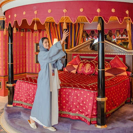 Woman in a blue dress standing next to a red bed with gold accents in a decorated room.