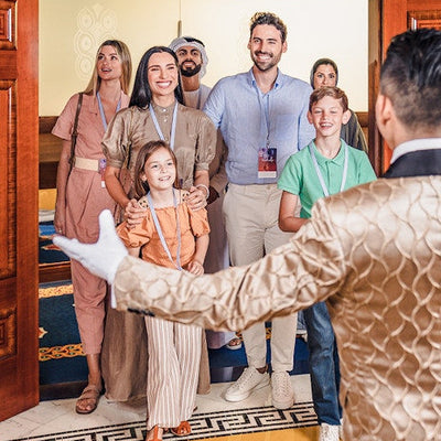 Group of people, including children, standing in a formal setting with a person in a patterned outfit extending a hand.