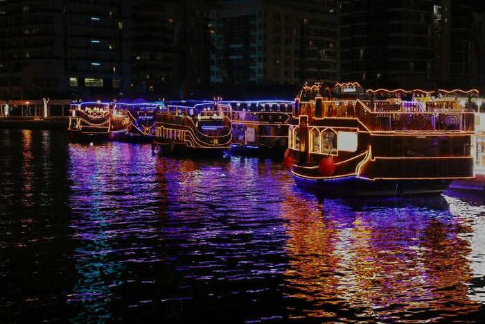 Promotional image for Dubai Marina Dhow Cruise Dinner featuring a vibrant nightscape of the Dubai Marina with a dhow boat, and textual information including the price and show details.