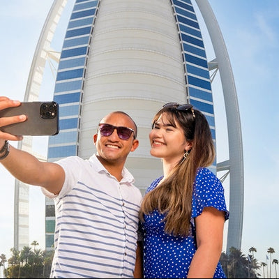 A man and a woman standing in front of the Burj Al Arab, with the man taking a picture of the woman with his smartphone.