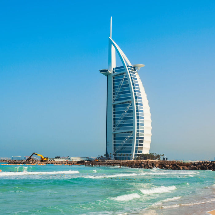 The image shows the Burj Al Arab hotel, with its distinctive sail-like design and blue facade, set against a clear blue sky and a calm sea background.